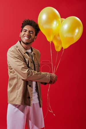 cheerful african american man in beige jacket holding helium balloons on red background, party