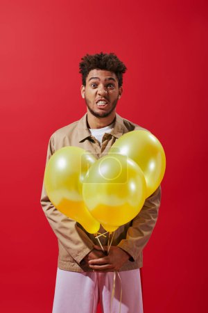 funny african american man in beige jacket holding balloons and grimacing on red background