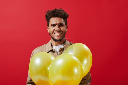 funny african american man in beige jacket holding balloons and grinning on red background
