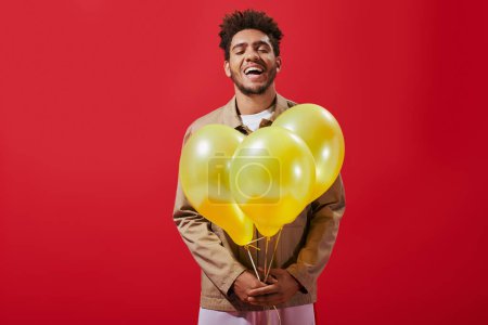 cheerful african american man in beige jacket holding balloons and laughing on red background