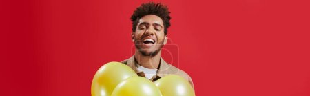 happy african american man in beige jacket holding balloons and laughing on red background, banner