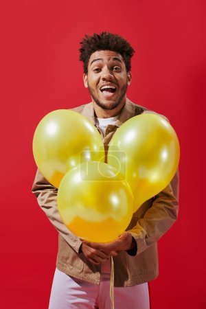 positive african american man in beige jacket holding balloons and laughing on red background