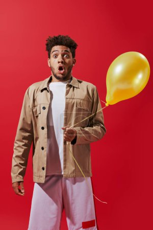 shocked african american man in beige jacket holding helium balloon on red background, party