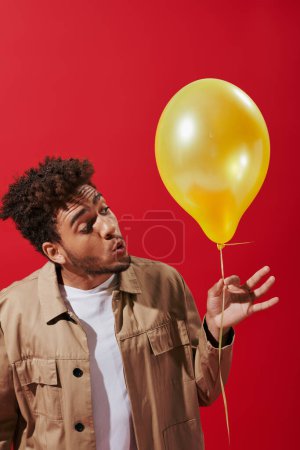 curly african american man in beige jacket looking at helium balloon on red background, party