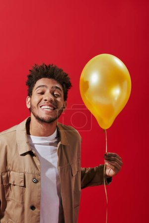 positive african american man in beige jacket holding balloon and smiling on red background