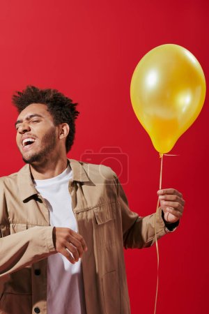 optimistic african american man in beige jacket holding balloon and smiling on red background magic mug #692588806