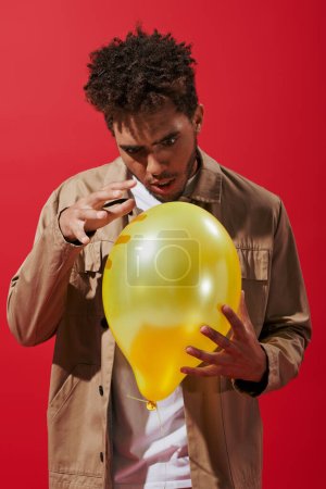 enchanted african american man in beige jacket looking at balloon on red background, amusement