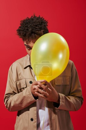 joven afroamericano hombre en chaqueta beige escondido detrás de globo sobre fondo rojo, oscureciendo la cara