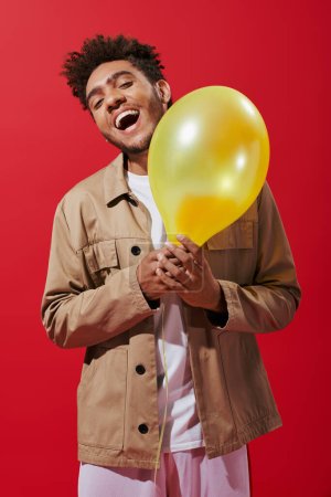 excited african american man in beige jacket holding balloon and smiling on red background