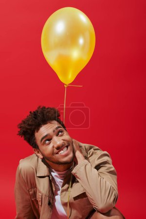 pleased african american man in beige jacket smiling near yellow balloon on red background