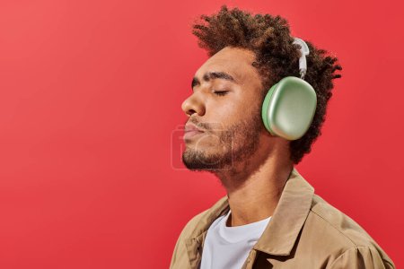 portrait of young african american man in wireless headphones listening music on red background