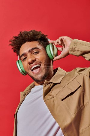 portrait of excited african american fella in wireless headphones listening music on red background
