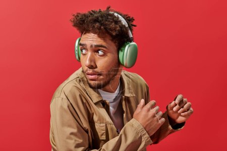 Photo for Portrait of confused african american man in wireless headphones looking away on red background - Royalty Free Image