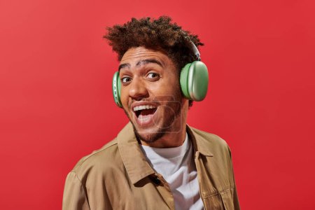 Porträt eines aufgeregten afrikanisch-amerikanischen Mannes in drahtlosen Kopfhörern mit offenem Mund auf rotem Hintergrund