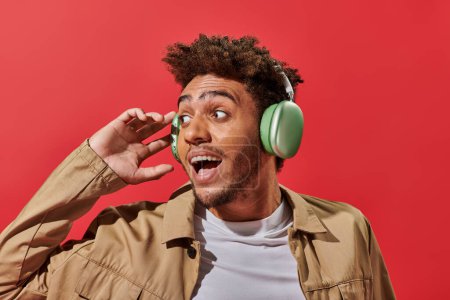 portrait de l'homme afro-américain étonné dans des écouteurs sans fil avec bouche ouverte sur fond rouge