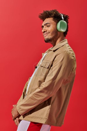retrato del hombre afroamericano complacido en auriculares inalámbricos disfrutando de música sobre fondo rojo