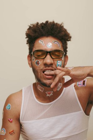 young african american man in sunglasses with trendy stickers on face biting finger on grey backdrop