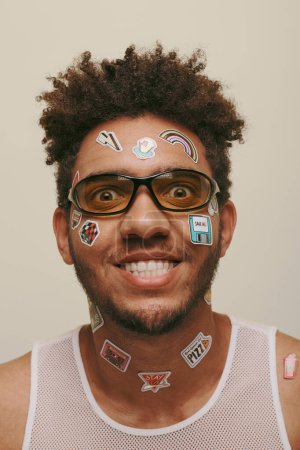 cheerful african american man in sunglasses with trendy stickers on face on grey background