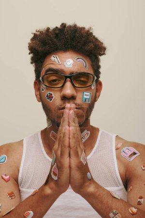 young african american man in sunglasses with trendy stickers on face and praying hands on grey