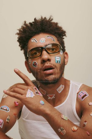 amazed young african american man in sunglasses with trendy stickers on face on grey background