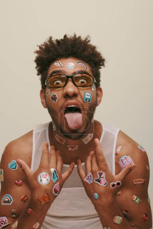 eccentric young african american man in sunglasses with trendy stickers on face on grey background