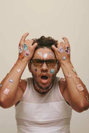 shocked young african american man in sunglasses with trendy stickers on face on grey background