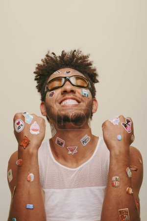 smiling young african american man in sunglasses with trendy stickers on face on grey background