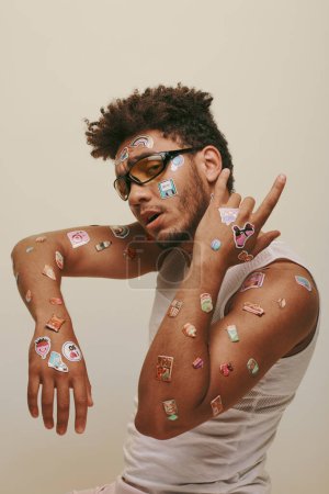 african american guy in sunglasses with stickers on face looking at camera on grey background