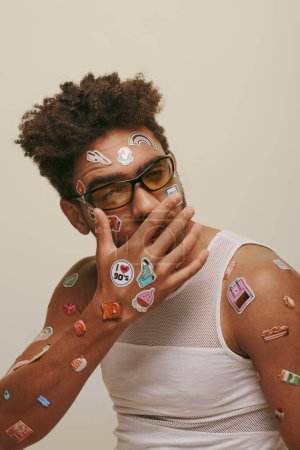 african american guy in sunglasses with stickers on face covering mouth on grey background