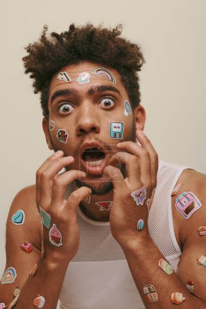 shocked african american guy in tank top with stickers on face looking at camera on grey background