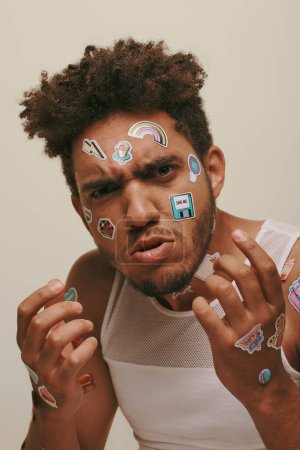 angry african american guy in tank top with stickers on face looking at camera on grey background