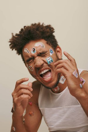 excited african american guy in tank top with stickers on face and closed eyes on grey background