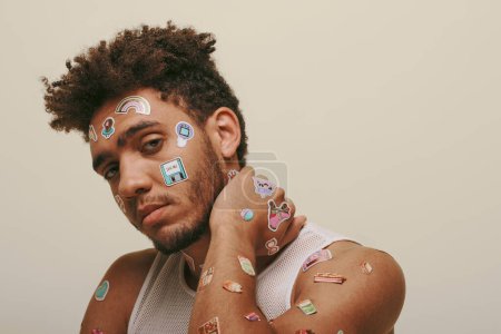 african american guy in tank top with stickers on face looking at camera on grey background