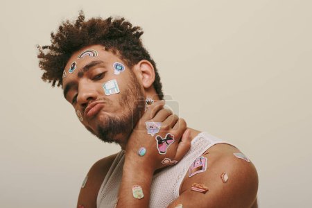 african american young man in tank top with stickers on face looking at camera on grey background