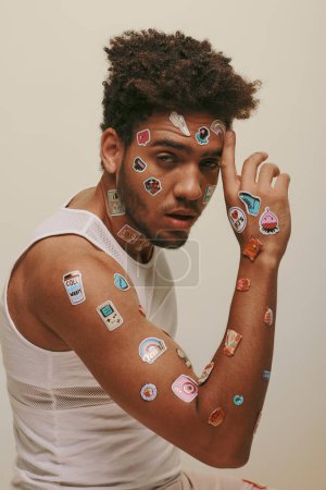 hombre afroamericano fresco en camiseta sin mangas con pegatinas en la cara mirando a la cámara sobre fondo gris