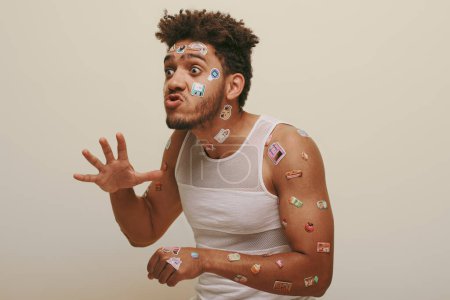 ausdrucksstarker afrikanisch-amerikanischer Mann mit Aufklebern auf dem Gesicht, Grimassen färben und gestikulieren auf grauem Hintergrund