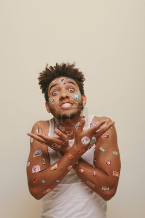 emotional african american man with stickers on face gesturing on grey background, hipster