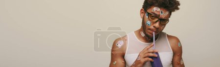 young african american guy with stickers on face and body drinking soda on grey background, banner