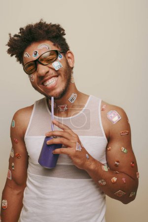 optimista afroamericano hombre con pegatinas en la cara y el cuerpo sosteniendo lata de refresco sobre fondo gris