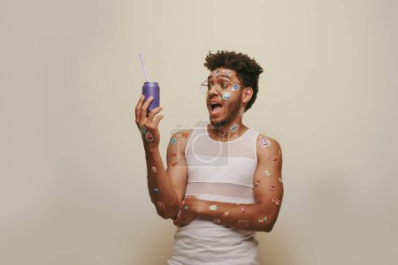 asombrado hombre afroamericano con pegatinas en la cara y el cuerpo mirando lata de refresco sobre fondo gris
