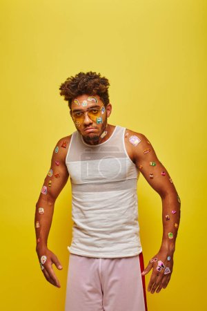 unzufriedener junger afrikanisch-amerikanischer Mann mit Aufklebern im Gesicht und Körper auf gelbem Hintergrund