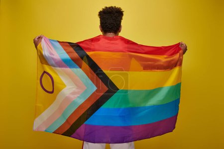 vista posterior del hombre afroamericano sosteniendo la bandera del arco iris lgbtq sobre fondo amarillo, mes de orgullo