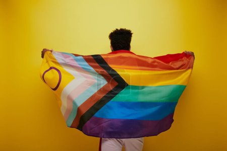 vista posterior del hombre afroamericano sosteniendo la bandera del arco iris lgbtq sobre fondo amarillo, mes de orgullo