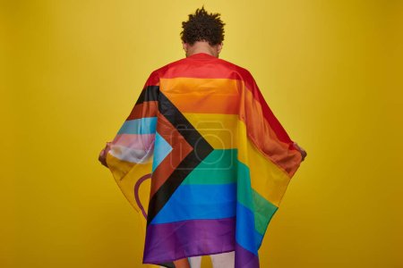 back view of african american man holding rainbow flag on yellow background, pride month