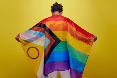 Foto de Vista posterior del chico afroamericano sosteniendo la bandera del arco iris lgbt sobre fondo amarillo, mes de orgullo - Imagen libre de derechos