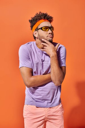 reflexivo joven afroamericano hombre en diadema y gafas de sol sobre fondo naranja, cara divertida