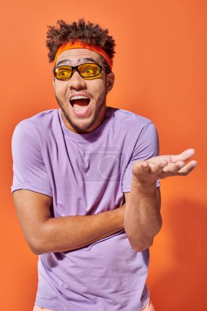 excited african american man in headband and sunglasses gesturing with open mouth on orange backdrop