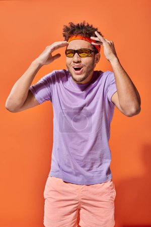 shocked african american man in headband and sunglasses gesturing on orange backdrop, mind blowing