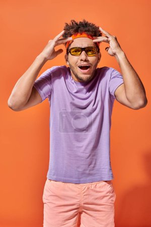 amazed african american man in headband and sunglasses gesturing on orange backdrop, mind blowing