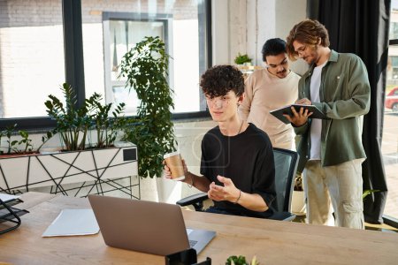 Curly-haired young man with coffee to go using laptop near male coworkers discussing project